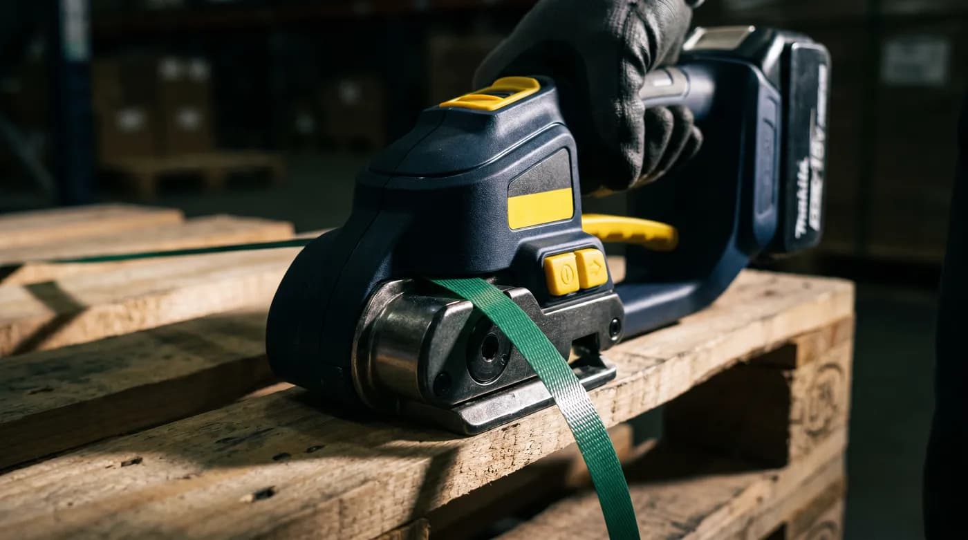 A cordless battery strapping tool tensioning plastic strap around a loaded pallet in a warehouse